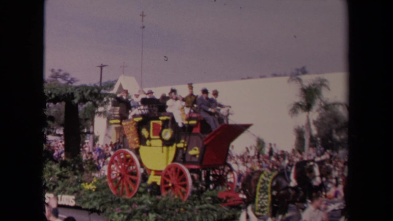 The stagecoach float moves through a sunny Los Angeles parade, with riders seated above red wheels and spectators behind it.