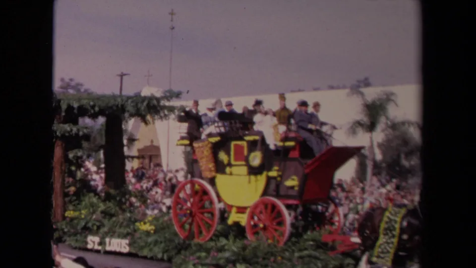 A stagecoach-themed parade float with yellow panels and red wheels passes a crowd, with a small sign near the curb reading St. Louis.