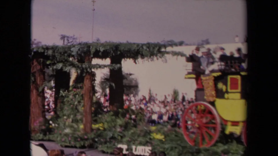 1967 Los Angeles parade view with a stagecoach float at right, floral arch structure, spectators, and partial route signage.