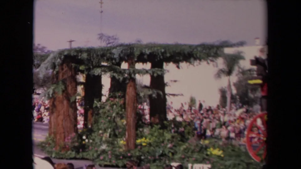 1967 Los Angeles parade frame showing a greenery-covered float structure, tree-like posts, flowers, spectators, and the edge of a red wheel.