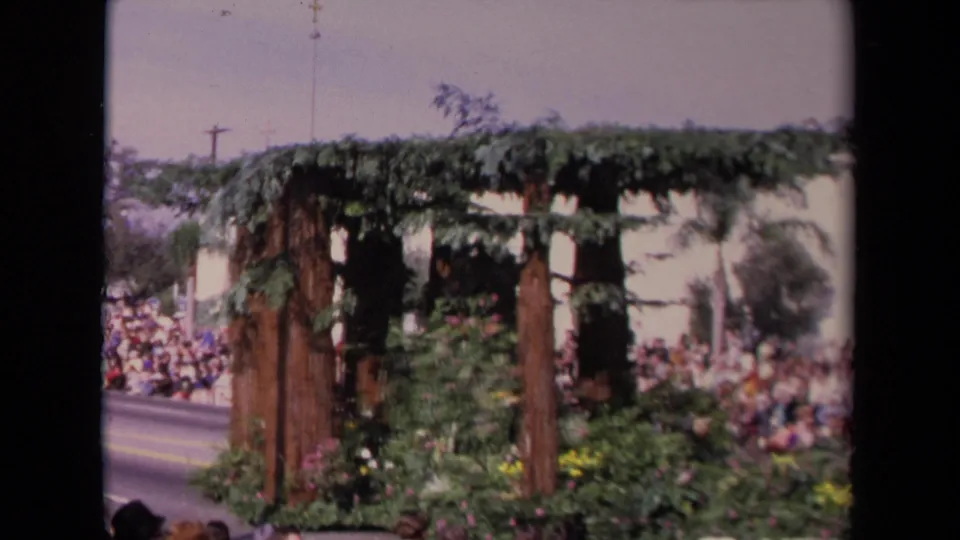 1967 Los Angeles parade view of a floral float section with upright wooded posts, dense greenery, flowers, and spectators lining the street.