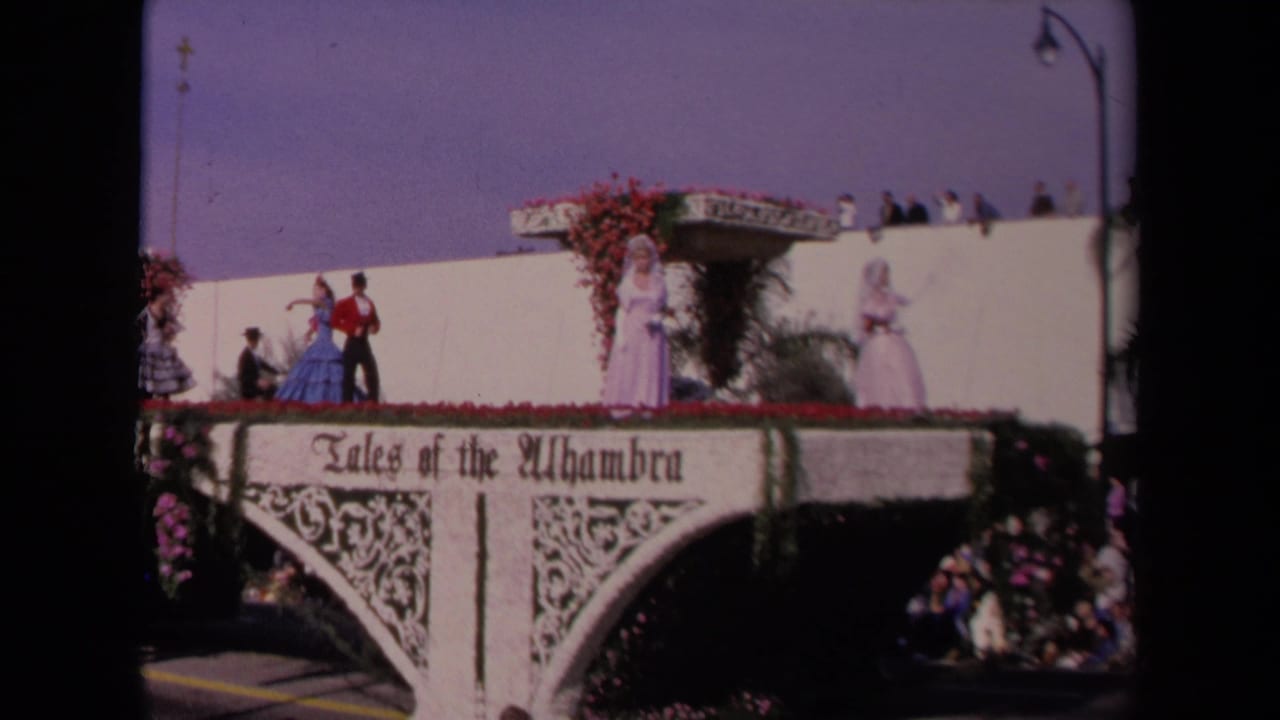 White parade float with Tales of the Alhambra lettering and costumed performers under a blue sky.