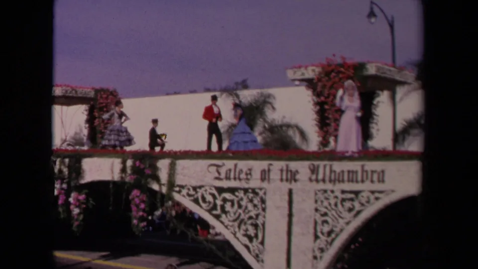 A 1967 Los Angeles home-movie view of the Tales of the Alhambra float, showing costumed performers beside floral arches and greenery.