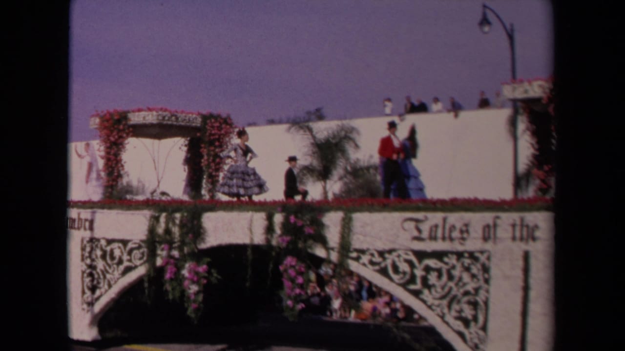 1967 Los Angeles home-movie frame showing Spanish-style costumed performers on a white flower-covered parade float with spectators above.