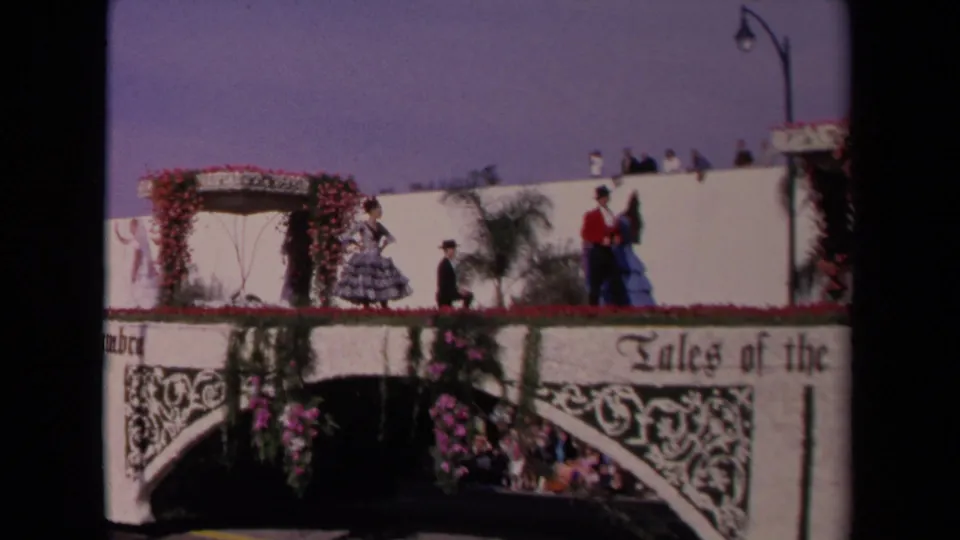 1967 Los Angeles home-movie frame showing Spanish-style costumed performers on a white flower-covered parade float with spectators above.