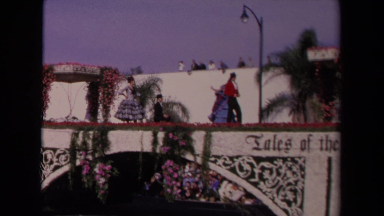 1967 Los Angeles 8mm frame of performers in Spanish-style costumes on a white floral float with partial "Tales of the" signage.