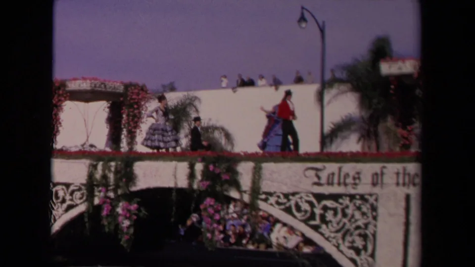 1967 Los Angeles 8mm frame of performers in Spanish-style costumes on a white floral float with partial "Tales of the" signage.