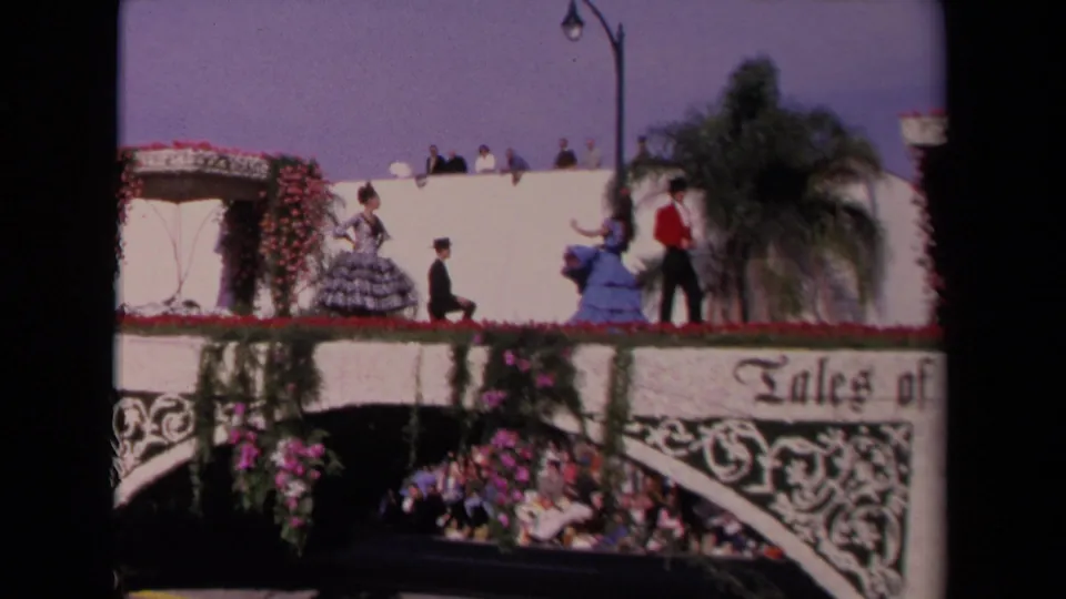 1967 Los Angeles parade frame showing costumed performers above ornate float trim, flowers, palm foliage, and seated spectators below.