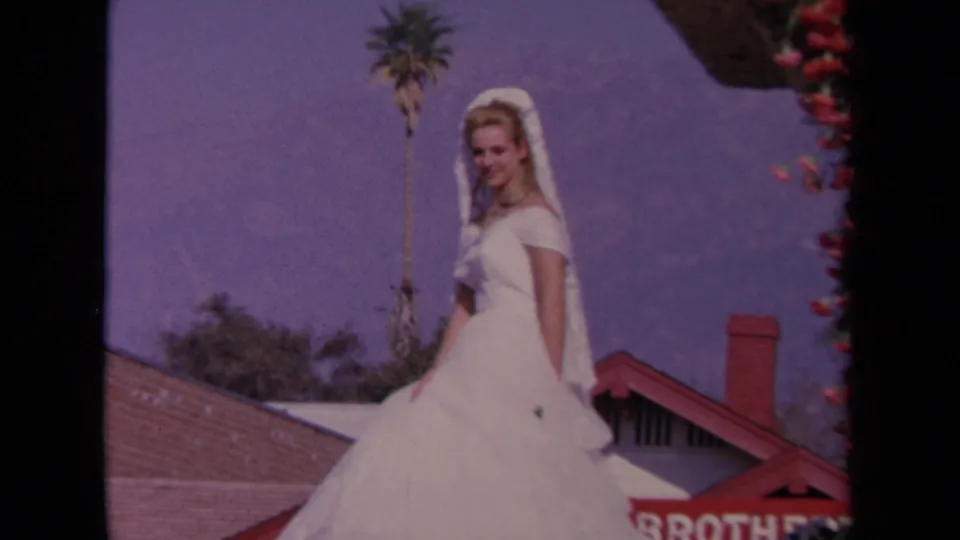 1967 Los Angeles home-movie frame showing a woman in a white costume on a parade float with palm tree and partial red lettering behind her.