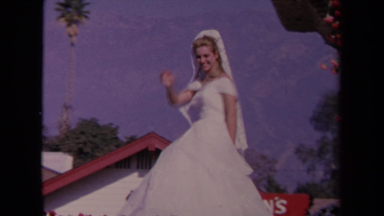 1967 Los Angeles 8mm frame of a bride in a white gown and veil waving from an outdoor ceremony setting with palm and mountains behind.