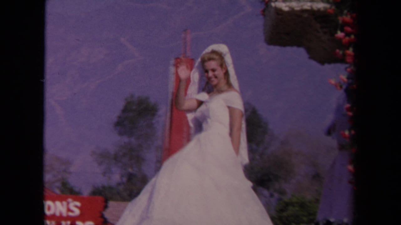 Bride in white waves beside floral trim with trees and mountains in the background.