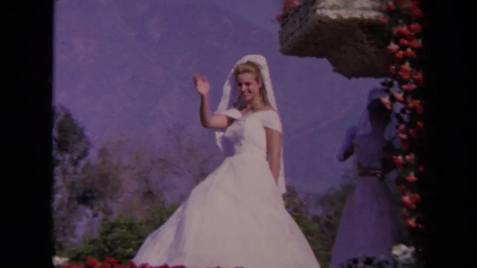 Soft 8mm frame from 1967 Los Angeles showing a waving bride in white, floral trim, mountains, shrubs, and another person partly visible nearby.