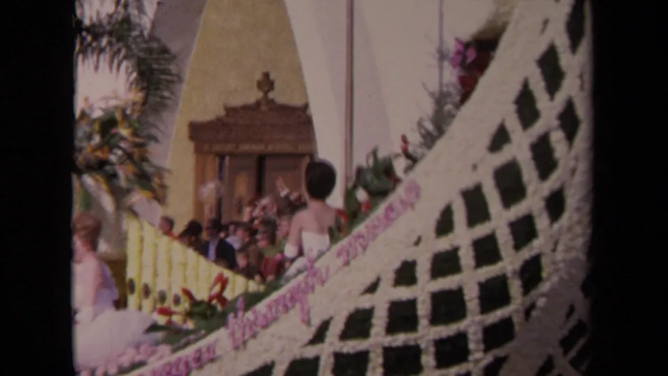 1967 Los Angeles 8mm frame showing guests, a church-like entrance, palm fronds, and a floral-decorated structure in a wedding or procession setting.
