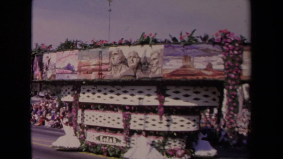 A 1967 Los Angeles parade float with painted scenic panels and flower trim moves past a crowd in bright daylight.