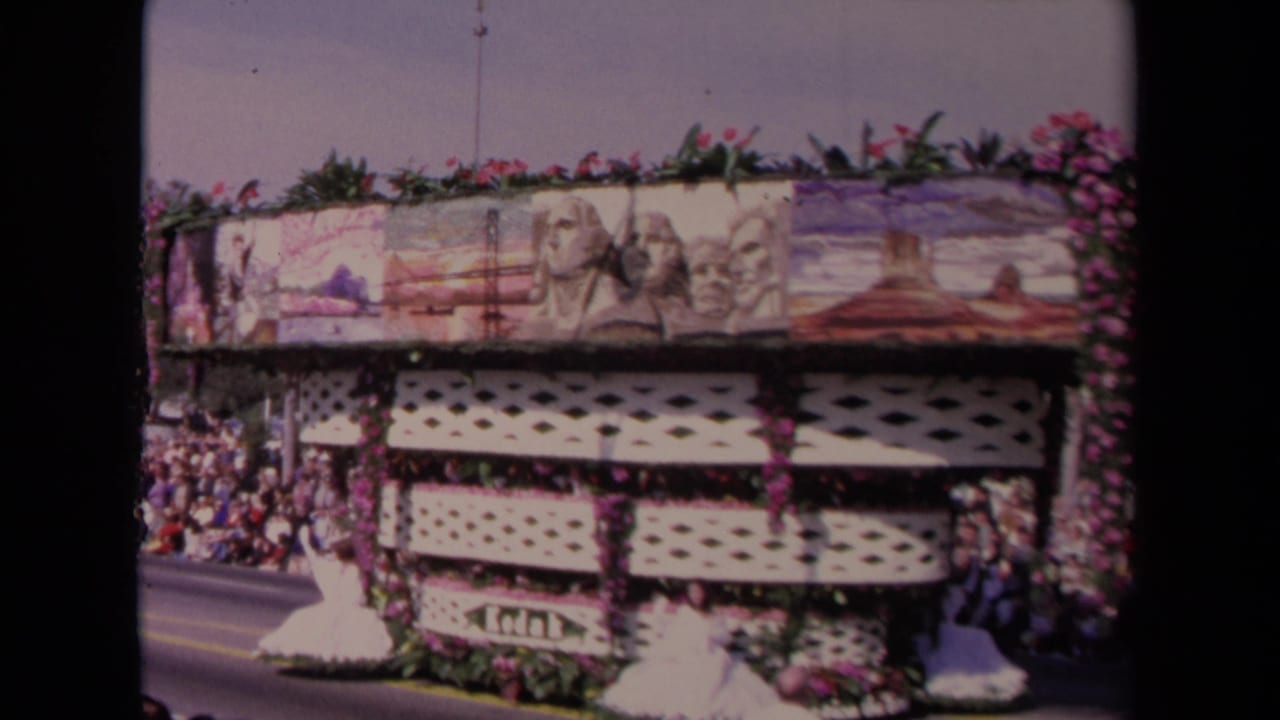 1967 Los Angeles parade float with painted national landmark panels, floral trim, crowd spectators, and a woman in white waving from the lower level.