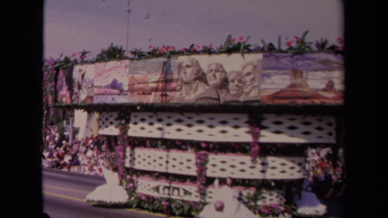 The 1967 Los Angeles parade float continues past the camera, showing floral trim, white lattice panels, landmark artwork, and spectators packed near the street.