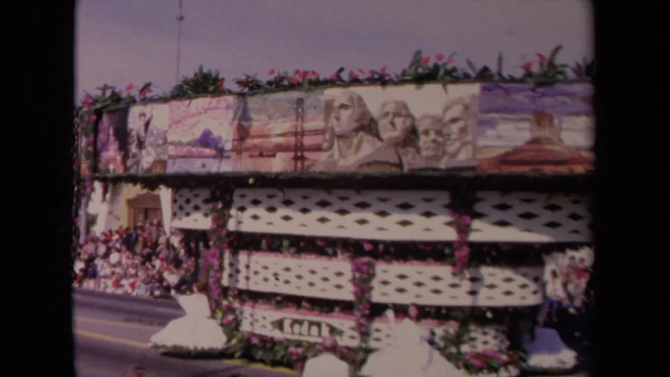 Side view of the 1967 Los Angeles parade float, with floral garlands, white latticework, painted landmark scenes, and spectators gathered beside the street.