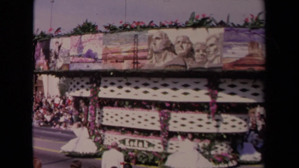 The large 1967 Los Angeles parade float passes close to camera, showing Mount Rushmore-style artwork, floral trim, lattice sides, and spectators along the route.