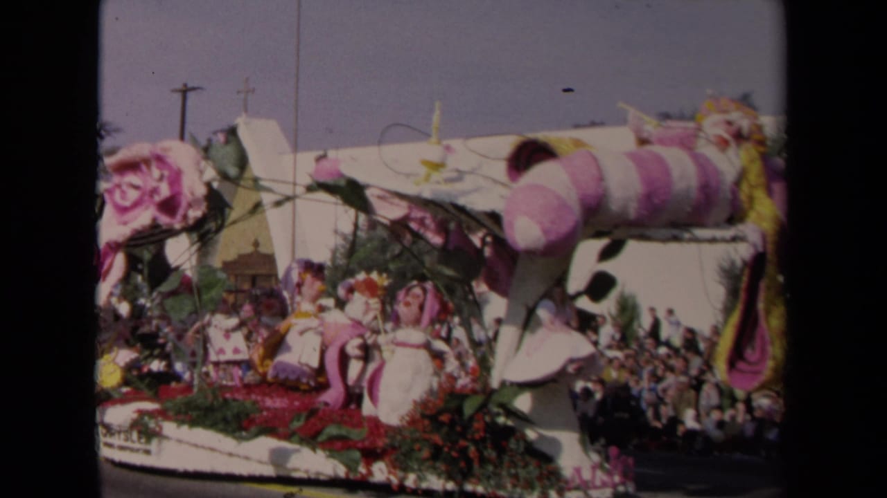 Parade float with large pink floral pieces and a crowd beside a white building.