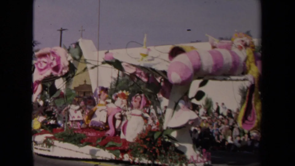 The float moves past a street crowd, with large pink decorations, greenery, and a white building visible in the background.