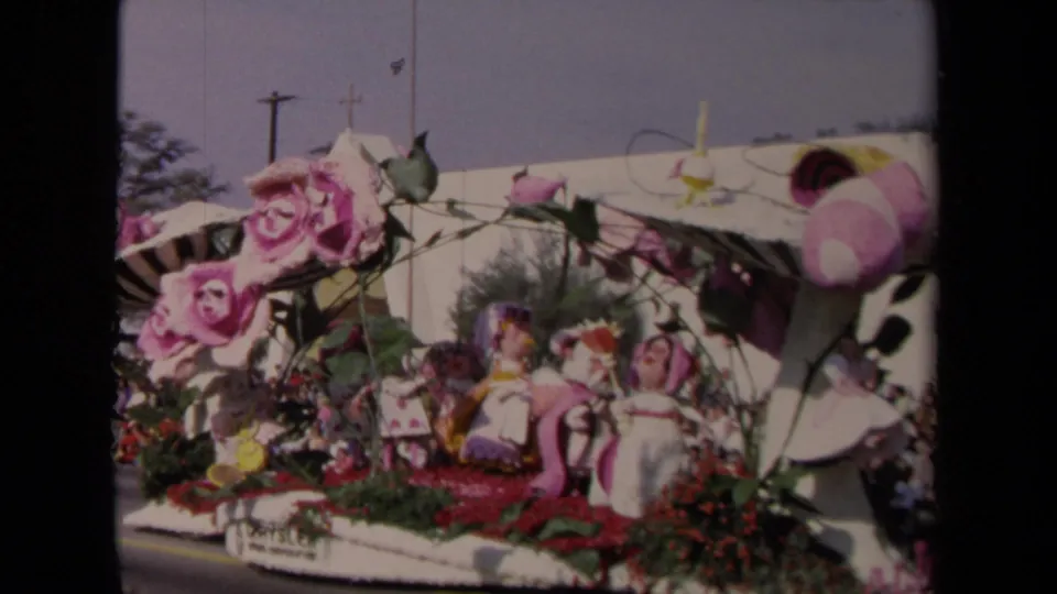 The final frame holds on the decorated parade float, showing large pink flowers, striped elements, figures, and a dense spectator crowd.