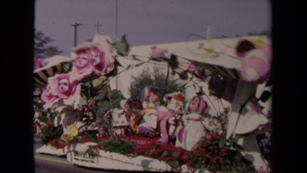 1967 Los Angeles home-movie frame showing a floral parade float with oversized pink roses, small figures, and a striped canopy.