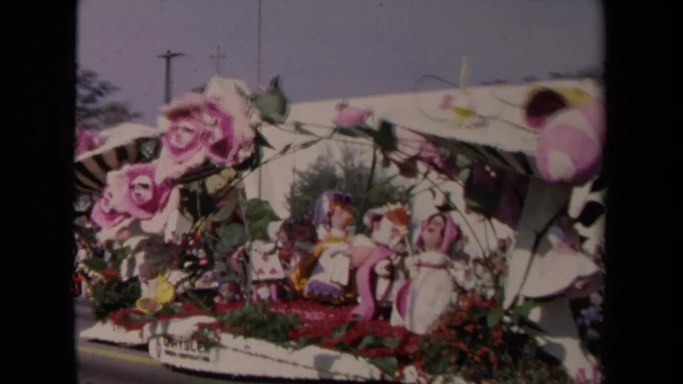 1967 Los Angeles home-movie frame showing a floral parade float with oversized pink roses, small figures, and a striped canopy.