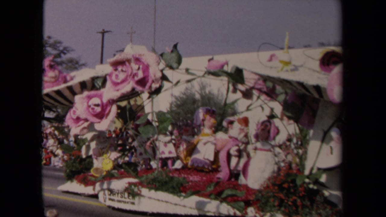 Floral parade float with large pink roses, canopy, and figure decorations viewed from the street.