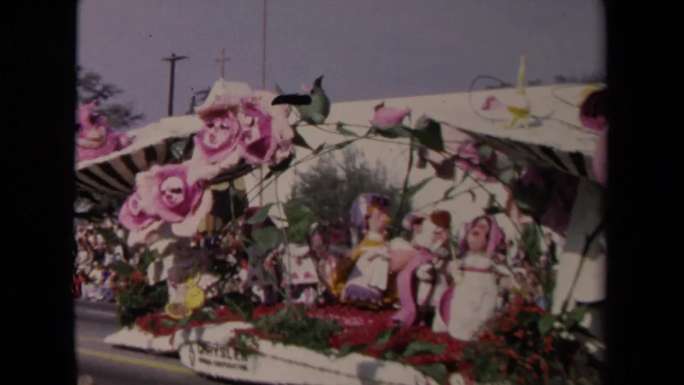 1967 Los Angeles 8mm frame showing a passing floral parade float with pink roses, figure displays, and dense red and green trim.