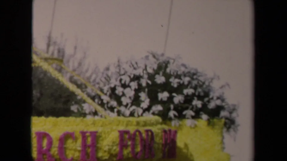 Close 1967 Los Angeles parade frame showing yellow floral float lettering with white blossoms and foliage behind it.