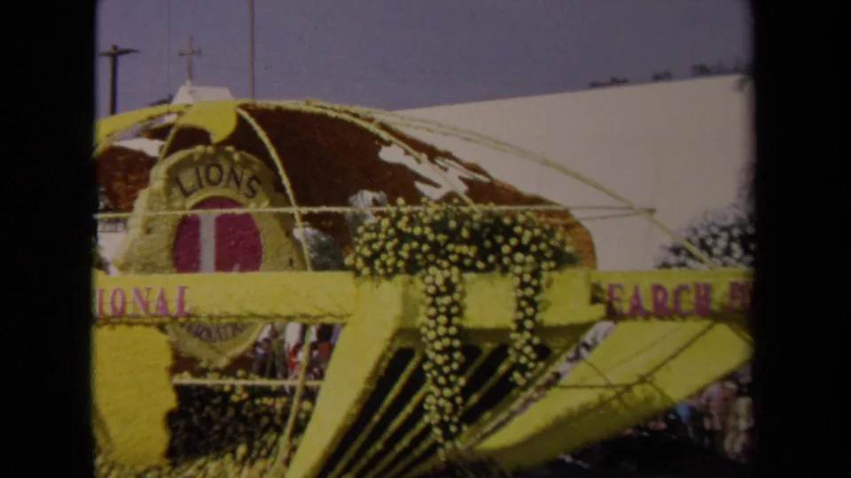 The Lions International parade float is seen more fully, with a yellow floral globe, purple text, and spectators lining the street below.