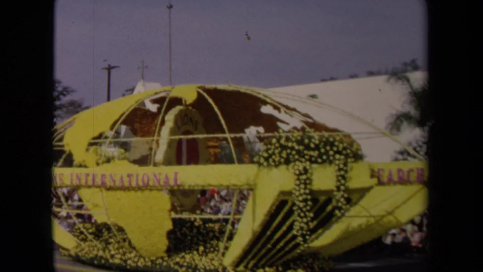 Wide 1967 Los Angeles street-parade view of the yellow Lions International globe float, purple lettering, and spectators beneath the float edge.