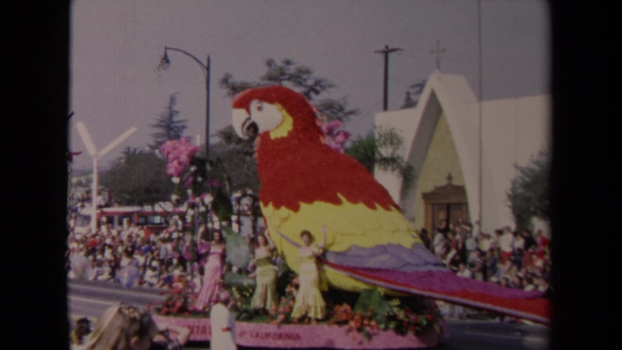 Giant floral parrot parade float with three women in gowns and spectators along the street.