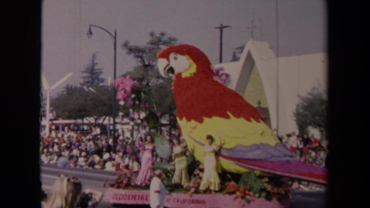 1967 Los Angeles parade view of a red and yellow floral parrot float with formally dressed women and spectators.