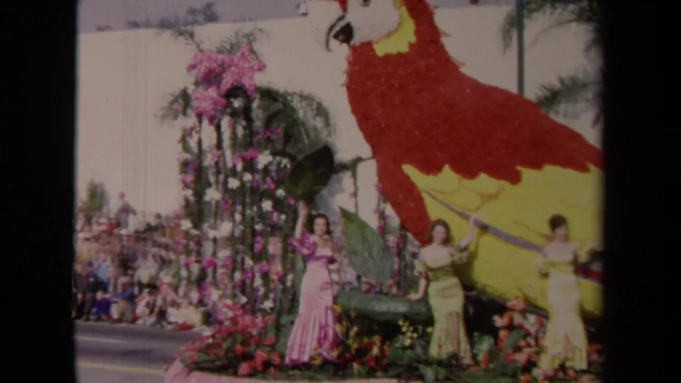 1967 Los Angeles parade frame with the floral parrot cropped at right, flower arch, two women, and spectators beyond.