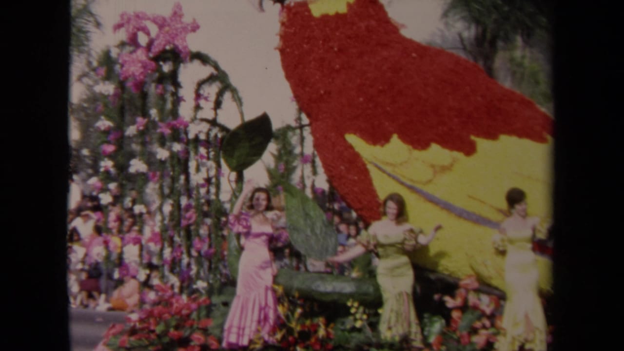 Three women wave from a floral parade float with a large red and yellow parrot figure.