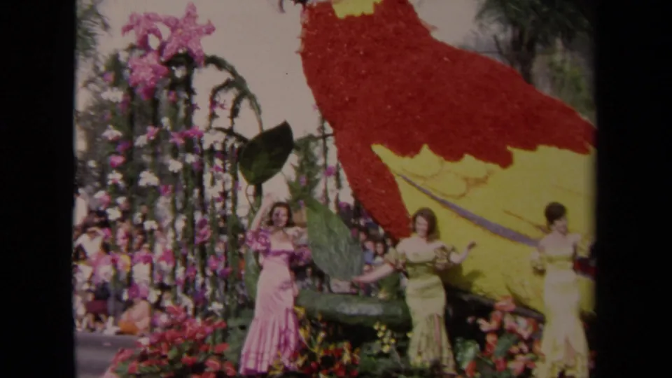 1967 Los Angeles home-movie frame showing women in pastel gowns waving beside a large floral parrot parade float.