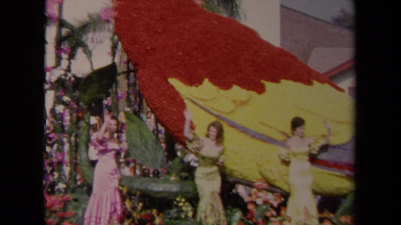 1967 Los Angeles parade frame with costumed women waving from a floral float built around a red and yellow parrot.
