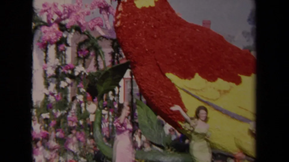 1967 Los Angeles home-movie frame with a woman waving beside the floral parrot figure on a parade float.