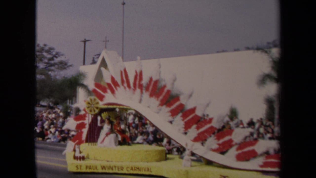 1967 Los Angeles parade float with a red-and-white feathered arch, yellow base, spectators, palm trees, and a church steeple in the background.