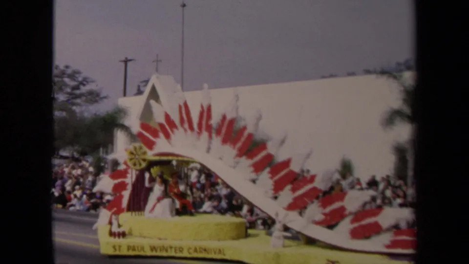 1967 Los Angeles parade float with a red-and-white feathered arch, yellow base, spectators, palm trees, and a church steeple in the background.