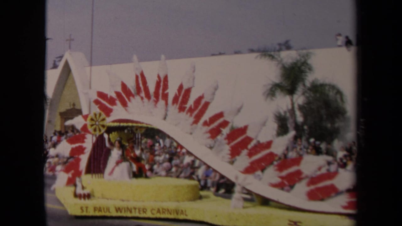 Decorated parade float with seated riders passes a large sidewalk crowd.