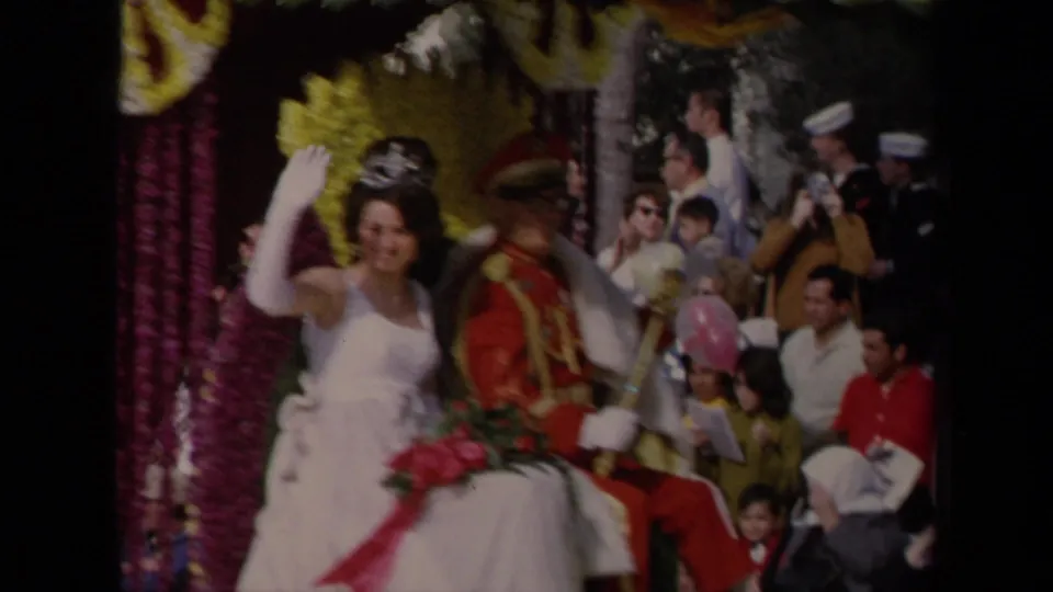 Closer 1967 parade view of a tiara-wearing woman in a white gown waving from the float beside a man in a red ceremonial uniform.