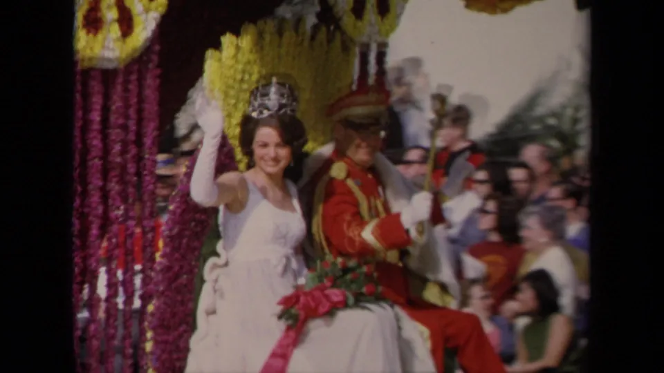 Close 1967 Los Angeles parade frame of the crowned woman waving from a floral float with a uniformed man and spectators behind her.