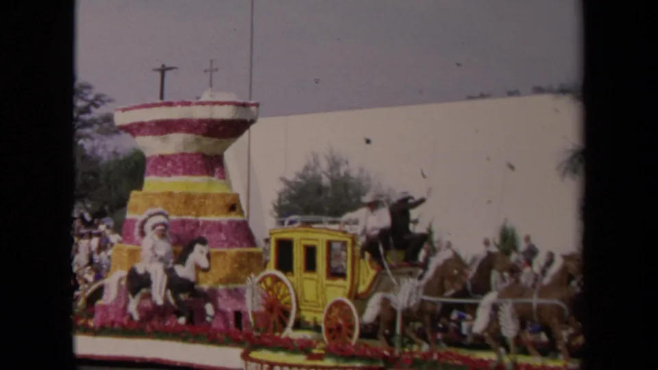 1967 Los Angeles parade float with a tiered floral structure, yellow stagecoach, horses, costumed riders, and crowds behind it.