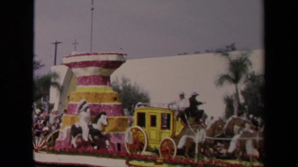 The Los Angeles parade float moves past spectators, showing a yellow stagecoach, horses, and a pink, white, and yellow floral tower.