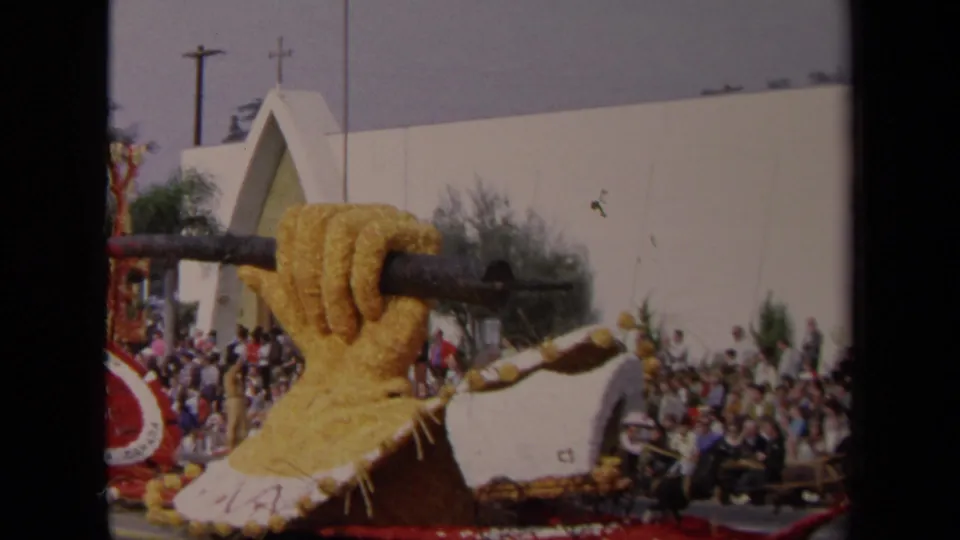 The parade float detail shows a large yellow floral hand around a rail, a horse figure, and the Los Angeles crowd in the background.