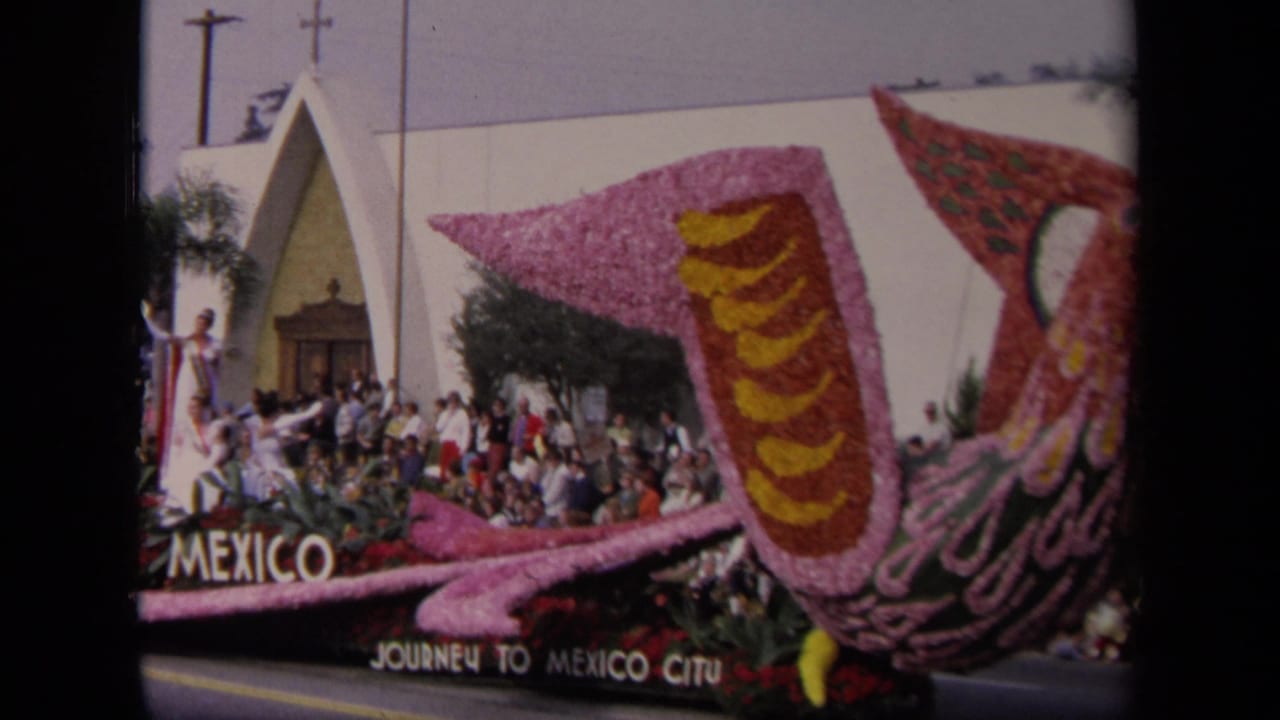 1967 Los Angeles parade frame showing a Mexico-themed floral bird float, spectators, palm trees, and a white modern building.