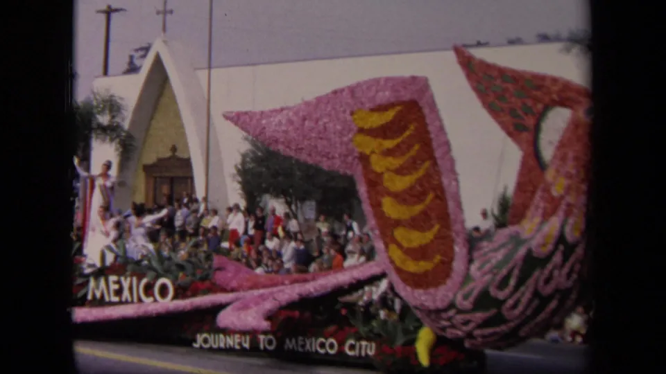 1967 Los Angeles parade frame showing a Mexico-themed floral bird float, spectators, palm trees, and a white modern building.