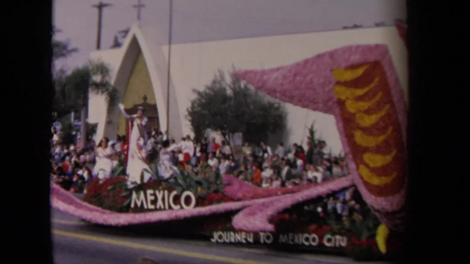 1967 Los Angeles frame of the Mexico float passing a crowd, with floral bird details, signage, palms, and a white building visible.
